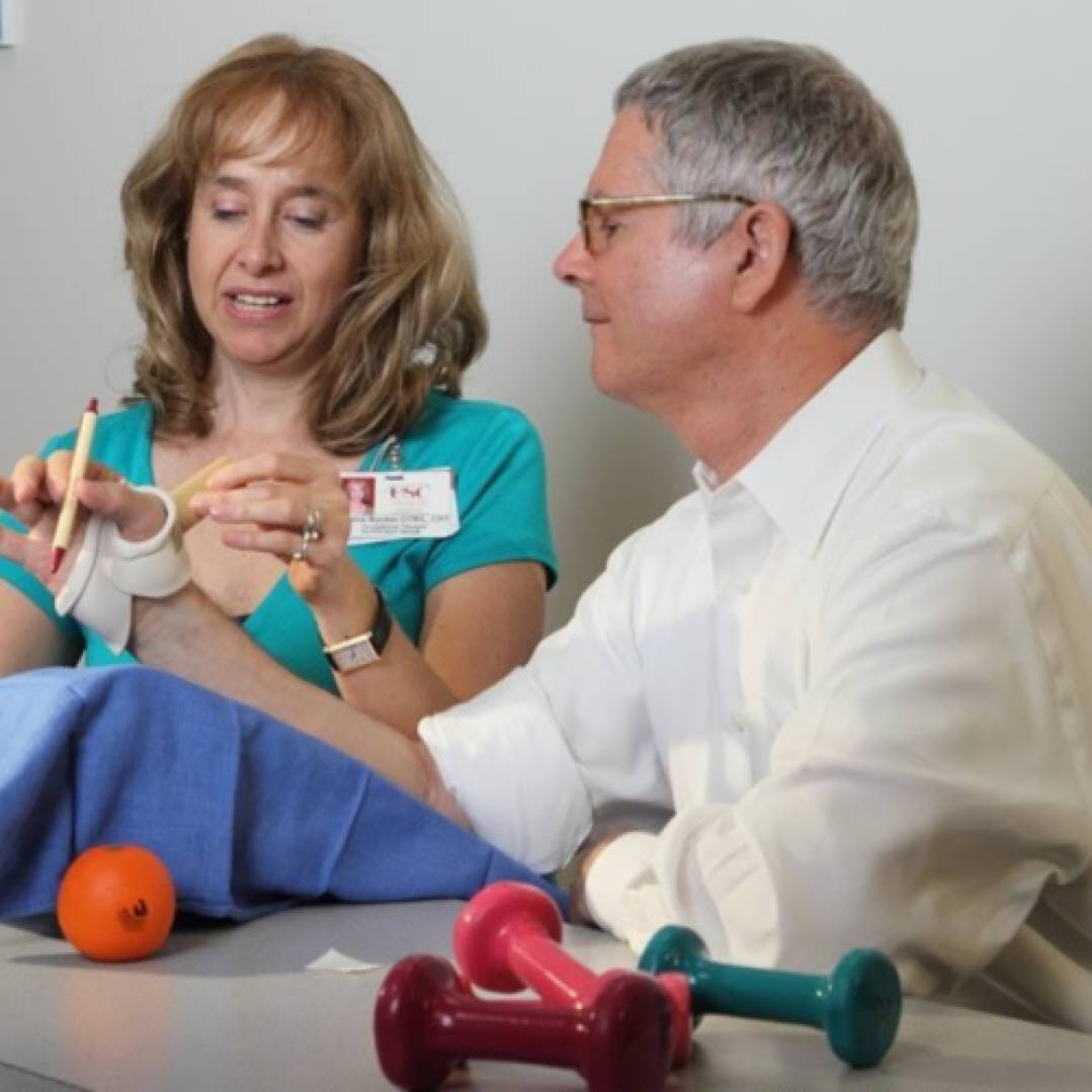 Occupational therapist working with an older patient. The patient holds a pen between thumb and forefinger while the therapist tightents a brace on his hand.